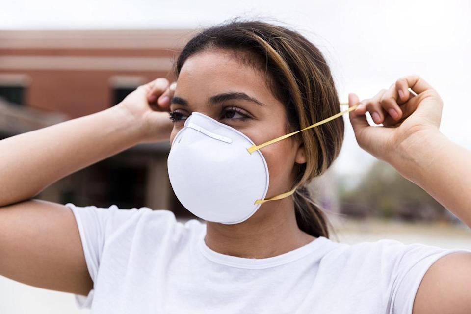 A mid adult woman protects herself by placing an N95 face mask over her nose and mouth.