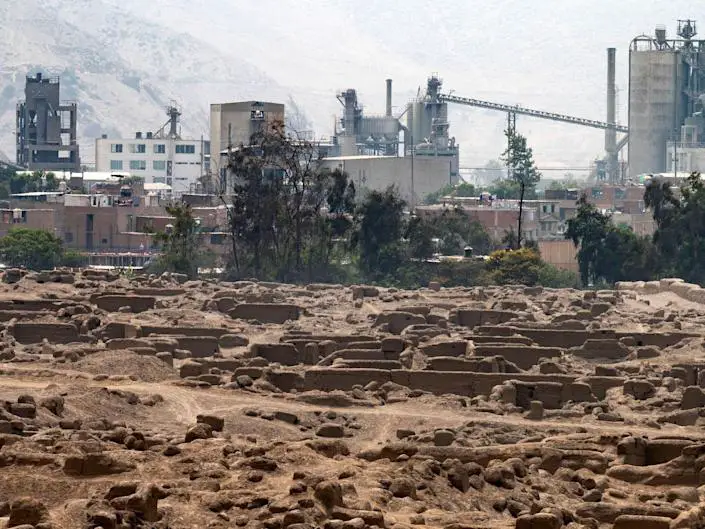A view of Cajamarquilla, an archeological site outside of Lima, Peru, shows the excavated landscape in front of a backgorund of industrial buildings.